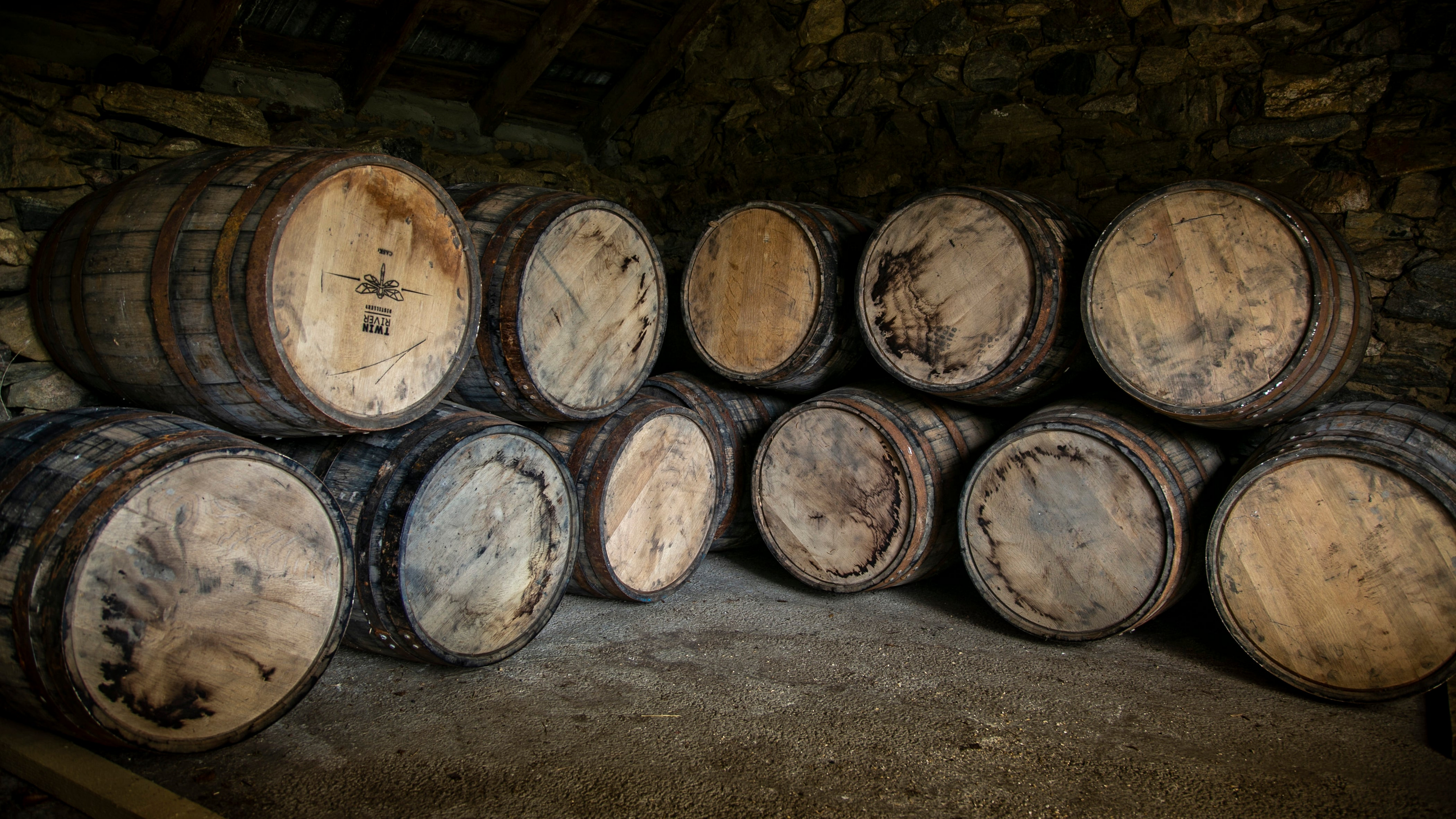 A row of large wooden barrels stacked inside a rustic stone cellar or warehouse with dim, warm lighting.